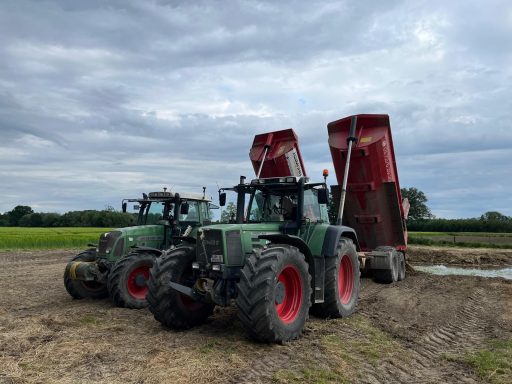 Zwei grüne Traktoren mit roten Anhängern auf einem Feld unter wolkigem Himmel.