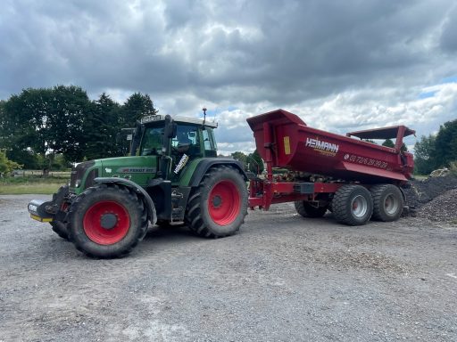 Traktor mit rotem Anhänger, der auf einem Kiesplatz steht, unter bewölktem Himmel.