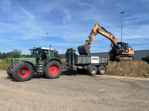 Traktor und Bagger laden Erde in einen Anhänger auf einem Baustellengelände.