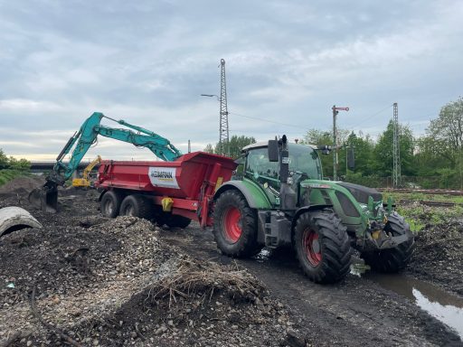 Traktor mit Anhänger und Bagger auf einem unebenen Feld bei bewölktem Himmel.